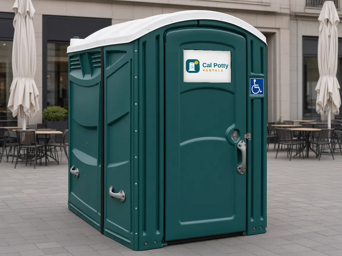 A teal ADA-accessible portable restroom with a wheelchair sign on a plaza patio with café tables and umbrellas